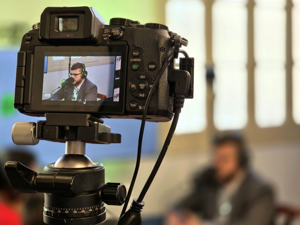 Ao fundo, parede branca com uma janela de batente bege e um homem sentado em uma cadeira, levemente desfocada. À frente, uma câmera digital fixada em tripé grava o podcast; no visor aparece um homem branco de cabelo e barba castanho-escuro, vestindo camisa social branca e terno, usando óculos e fone de ouvido preto com detalhes verdes. À sua frente há um microfone de podcast.