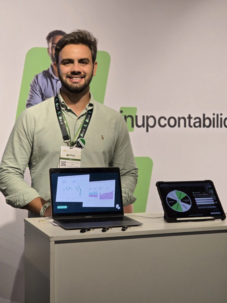 Ao fundo, banner branco com letras pretas e detalhes verdes da InUP Contabilidade. À frente, homem de barba e cabelo castanho-escuro, sorrindo, veste camisa social verde-clara e crachá do evento no pescoço. Ele apoia as mãos em um balcão branco, sobre o qual há um computador mostrando um slide de apresentação da InUP com chaveiros pretos à esquerda e um tablet com uma roleta de prêmios à direita.