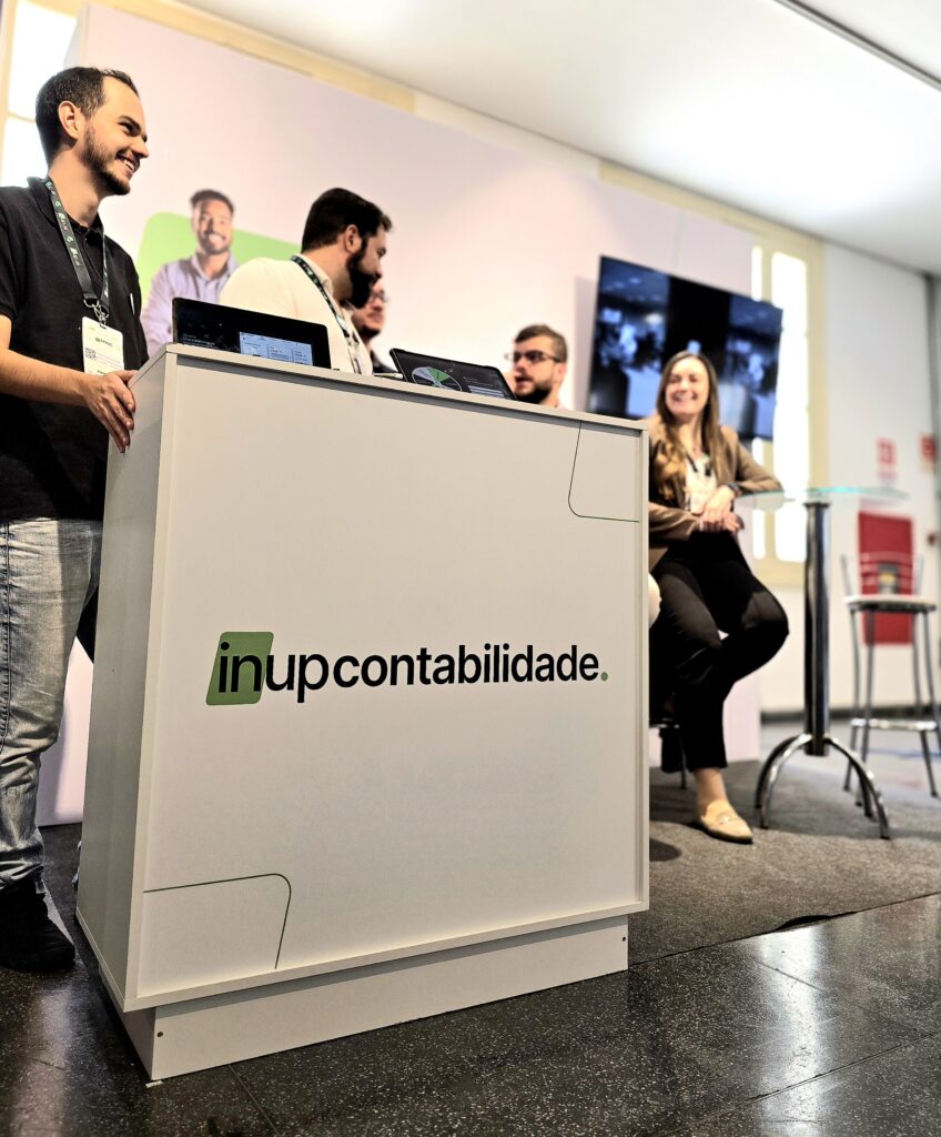 Ao fundo, banner branco com letras pretas e detalhes verdes da InUP Contabilidade. À frente, cinco pessoas: À esquerda, homem branco de cabelo e barba castanho-escuro, vestindo camisa preta, calça jeans clara, tênis preto e crachá do evento, sorrindo e olhando para a direita, com as mãos apoiadas em um balcão branco com o nome “InUP Contabilidade” em preto e detalhes verdes. Ao lado, homem branco de cabelo e barba pretos, camisa social branca e crachá do evento, olhando para a direita. Próximo a ele, homem branco de cabelo castanho usando óculos de grau. Em seguida, homem branco de cabelo e barba castanho-escuro, olhando para a esquerda. Por fim, mulher de cabelos longos castanho-claro, vestindo camisa social branca, blazer marrom-claro, calça social preta e sapato social bege, com o braço direito apoiado em uma mesa de vidro redonda, sentada em uma banqueta.