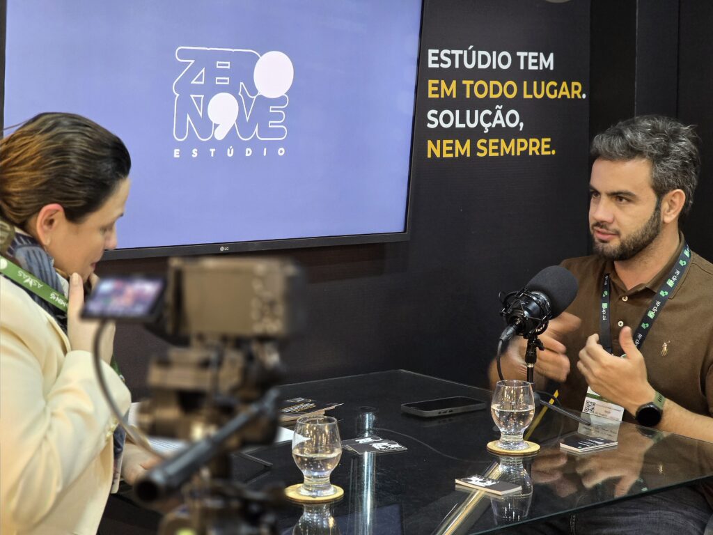 Ao fundo, parede preta com uma TV ao centro exibindo a logo do Estúdio Zero Novo. Ao lado, está escrito em branco e amarelo: “Estúdio tem em todo lugar. Solução, nem sempre”. À frente, uma mesa de vidro com dois participantes: À esquerda, mulher branca de cabelos castanhos presos, vestindo casaco bege e cachecol verde, azul e branco. À sua frente, um copo de água pela metade e, ao lado, uma câmera digital filmando o podcast, apontada para a direita, registrando o outro participante. À direita, homem branco de cabelos e barba castanho-escuro, vestindo camisa polo marrom, crachá do evento no pescoço e relógio no braço esquerdo. À sua frente, microfone de podcast e, à direita, seu celular.