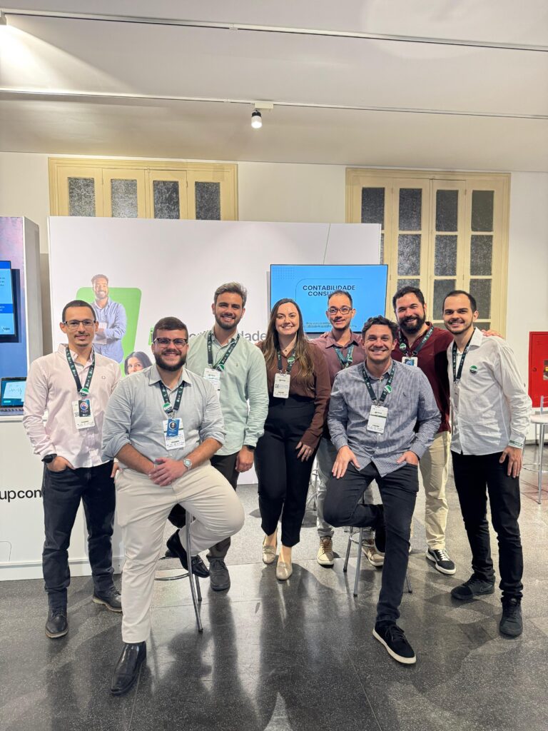 Ao fundo, parede branca com duas janelas de batente bege, banner branco da InUP Contabilidade e uma TV à direita exibindo uma apresentação sobre a empresa. À frente, o time InUP está reunido posando para uma foto.