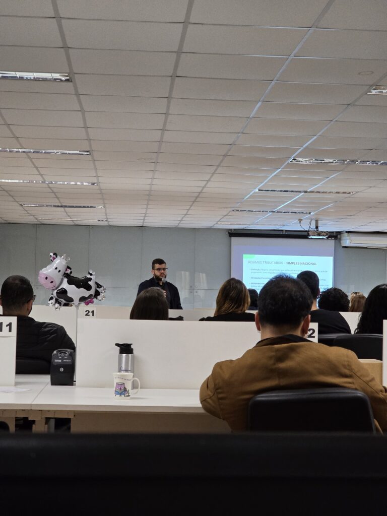 Ao fundo, um homem branco, de barba e cabelos castanhos, veste camisa social cinza-escuro e terno preto. Ele segura um microfone na mão direita enquanto apresenta um slide sobre os regimes tributários. O slide é branco, com texto em preto e detalhes em verde. Em primeiro plano, vê-se o público sentado de costas para a câmera, atento à apresentação.