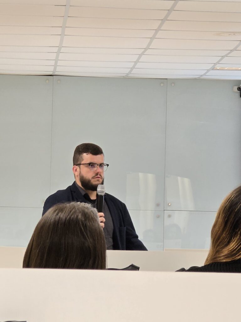 Ao fundo, um homem branco, de barba e cabelos castanhos, veste camisa social cinza-escuro e terno preto. Ele segura um microfone na mão direita.