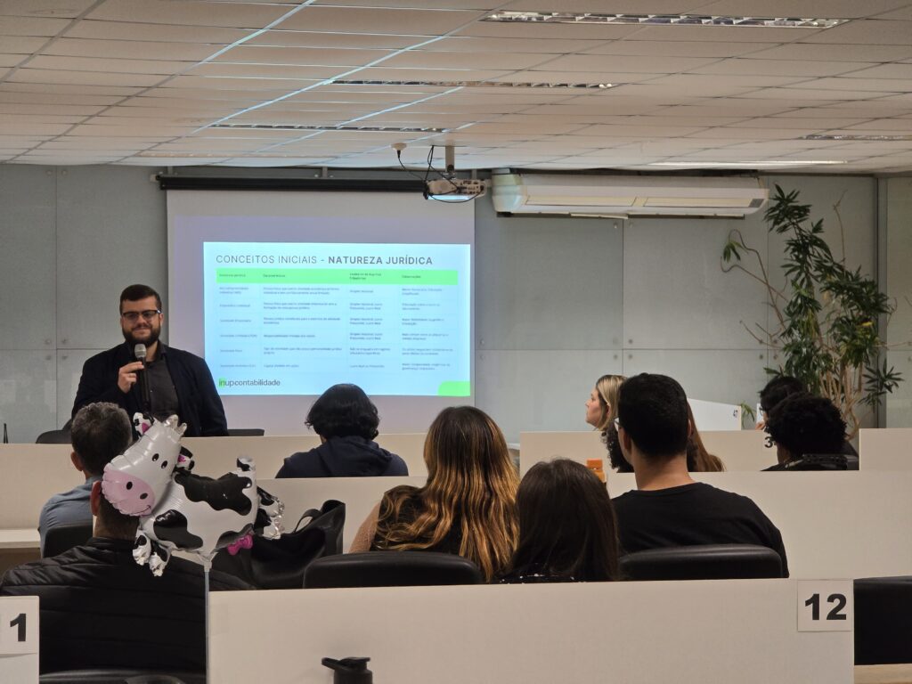 Ao fundo, um homem branco, de barba e cabelos castanhos, veste camisa social cinza-escuro e terno preto. Ele segura um microfone na mão direita enquanto apresenta um slide sobre os conceitos iniciais da natureza jurídica. O slide é branco, com texto em preto e detalhes em verde. Em primeiro plano, vê-se o público sentado de costas para a câmera, atento à apresentação.