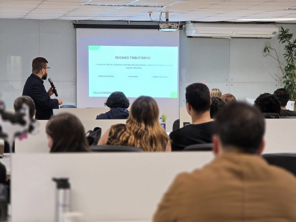 Ao fundo, um homem branco, de barba e cabelos castanhos, veste camisa social cinza-escuro e terno preto. Ele segura um microfone na mão direita e olha para um slide projetado sobre regimes tributários. O slide é branco, com texto em preto e detalhes em verde. Em primeiro plano, o público aparece de costas para a câmera, atento à apresentação.