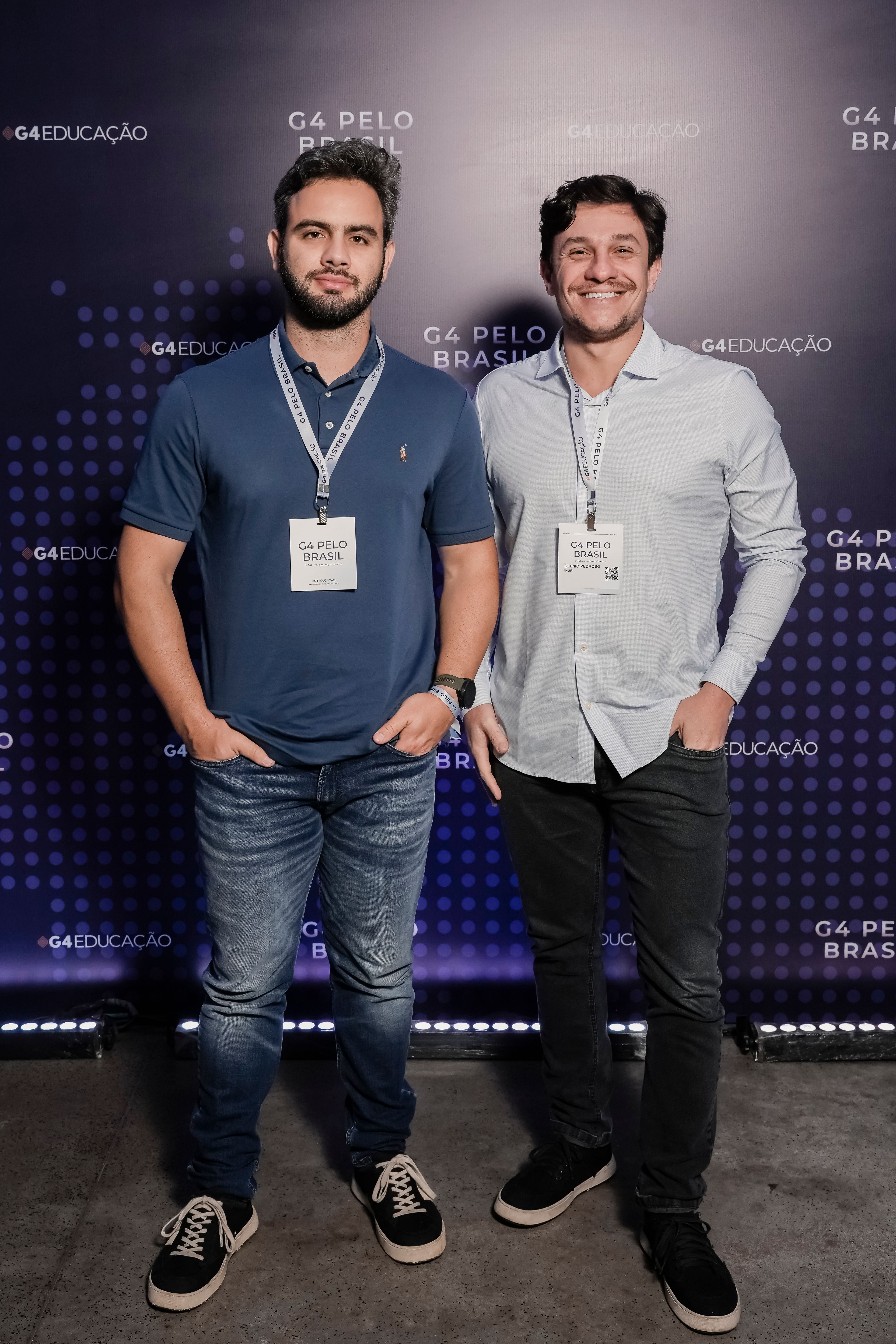 Dois homens brancos, sorrindo, com as mãos nos bolsos da calça. À esquerda, veste camisa polo azul-escura com logo amarela, calça jeans clara, relógio e pulseira do G4 Pelo Brasil no braço esquerdo e crachá do evento pendurado no pescoço. À direita, veste camisa social branca, calça jeans preta e crachá do evento pendurado no pescoço.