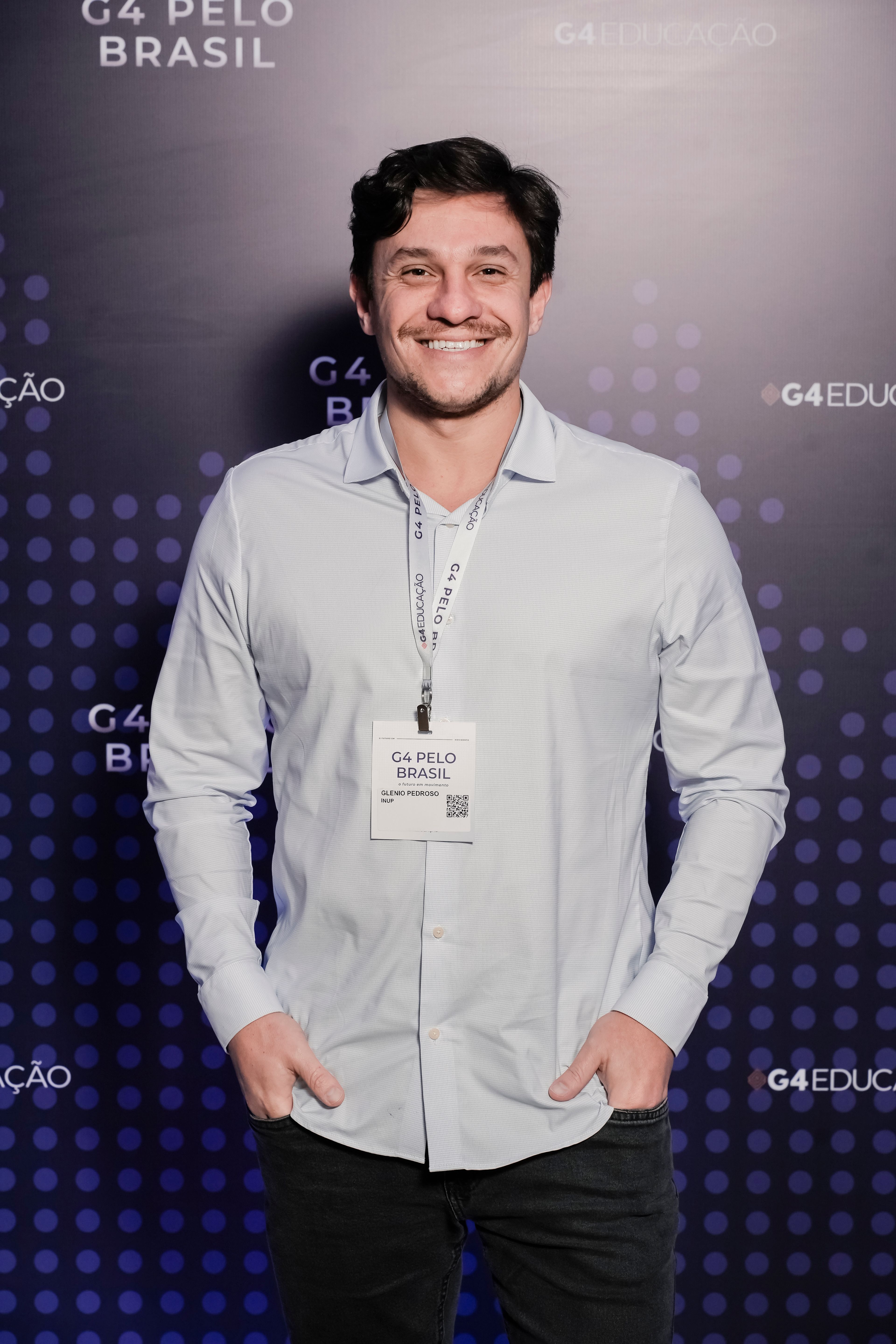Homem branco, de barba e cabelos castanho-escuro, sorrindo, com as mãos nos bolsos da calça. Veste uma camisa social branca, calça jeans preta e crachá do evento pendurado no pescoço.