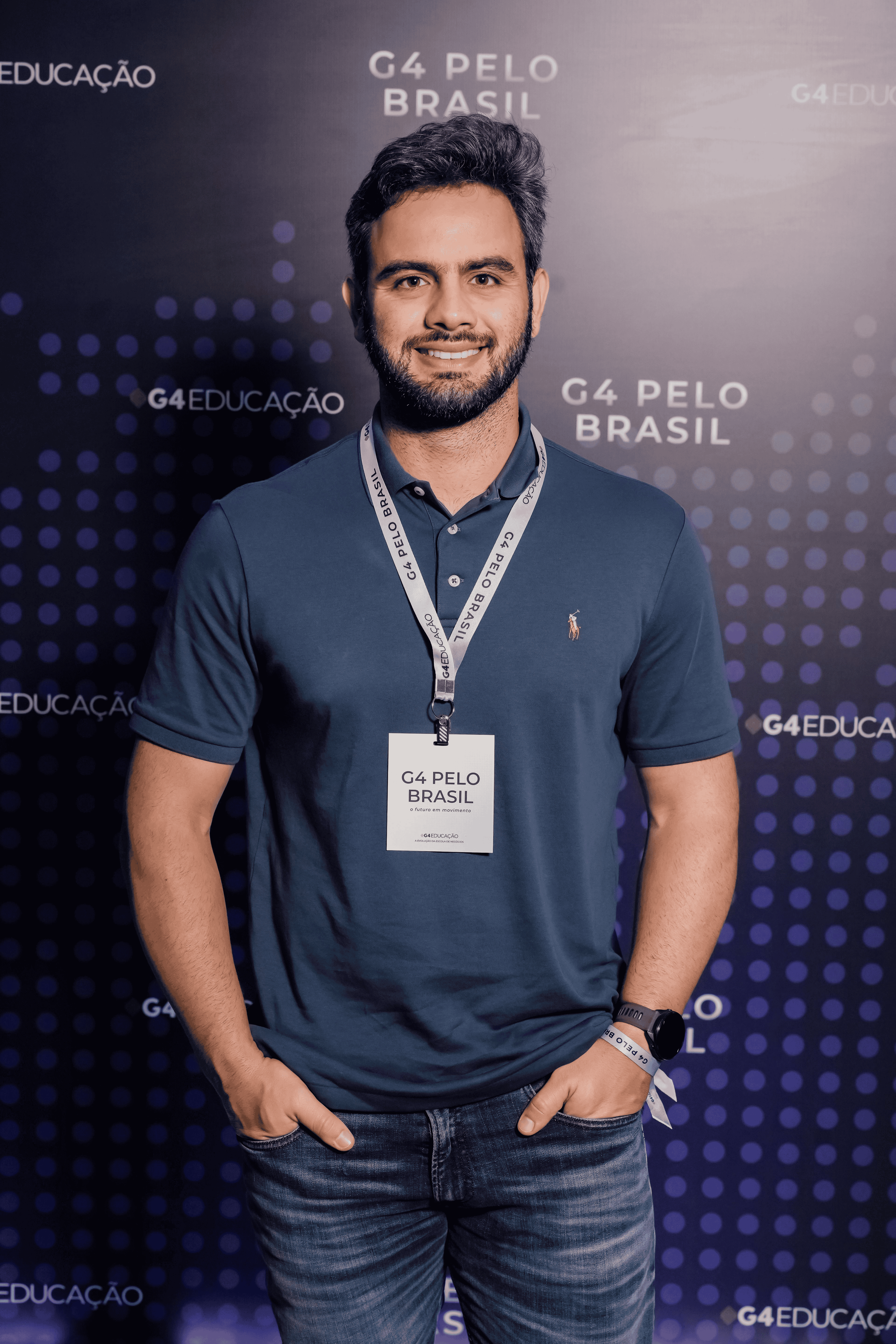 Homem branco, de barba e cabelos castanho-escuro, sorrindo, com as mãos nos bolsos da calça. Veste camisa polo azul-escura com logo amarela, calça jeans clara, relógio e pulseira do G4 Pelo Brasil no braço esquerdo, e crachá do evento pendurado no pescoço.