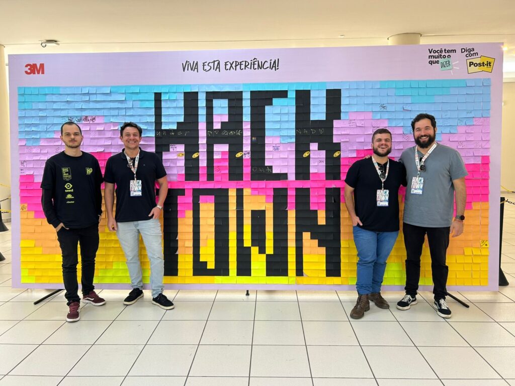 Painel de fundo coberto por post-its coloridos, em degradê do azul ao amarelo. No centro, a palavra “HackTown” formada com post-its pretos. No canto superior esquerdo está a logo da 3M, no centro a frase “Viva esta experiência!” e no canto superior direito a frase “Você tem muito o que dizer, diga com post-it”.
À frente do painel estão quatro homens, dois de cada lado, deixando o nome “HackTown” visível.
À esquerda: o primeiro veste calça preta, camisa preta de manga longa da InUP com detalhes verde-limão e tênis marsala com faixa branca; ao lado dele, outro homem de camisa preta, calça jeans clara, tênis preto e crachá do evento.
À direita: o primeiro usa camisa preta, calça jeans, sapato social marrom e crachá do evento; ao lado dele, o último veste camisa cinza, calça jeans, tênis preto e também crachá do evento.